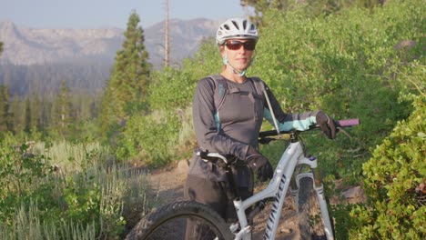 a mountain biker stands with her bike on a path near a forest