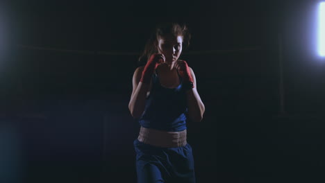 Professional-beautiful-female-boxer-otbryvatyvat-blows-conducting-a-fight-with-a-shadow-in-a-dark-hall-of-the-hall-in-slow-motion-in-blue-clothes-and-red-bandages-on-her-wrists.-flying-around-the-object.-steadicam-shot