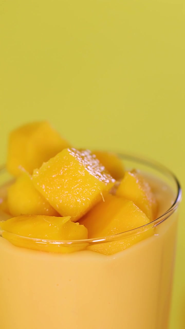 Primo piano di una mano che guarnisce un cremoso frullato al mango con cubetti di frutta e semi, su sfondo luminoso
