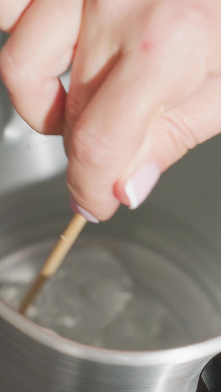 Close up of hand stirring liquid in metal container with wooden stick. Hand holds and moves stirrer through clear fluid creating ripples. Soft lighting highlights metallic bowl edges