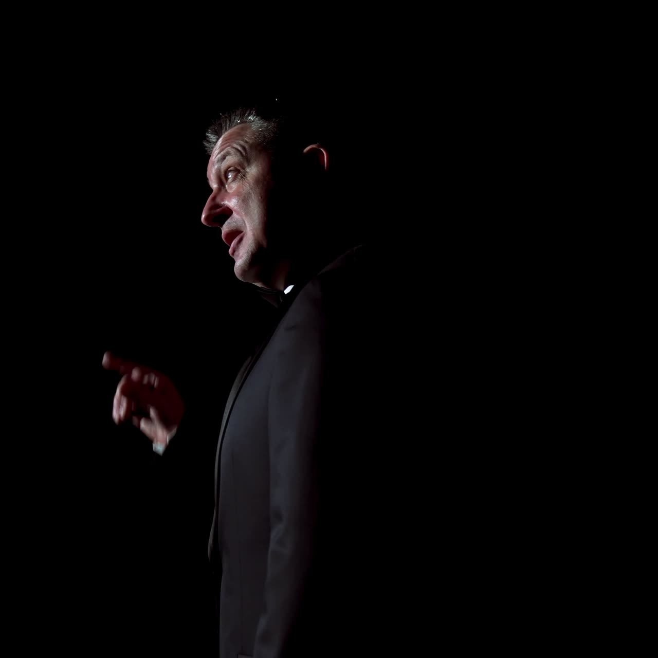 Male host talking on stage. Performer man in black suit standing on dark stage at spotlight and speaking with gestures in the theater. Circling camera