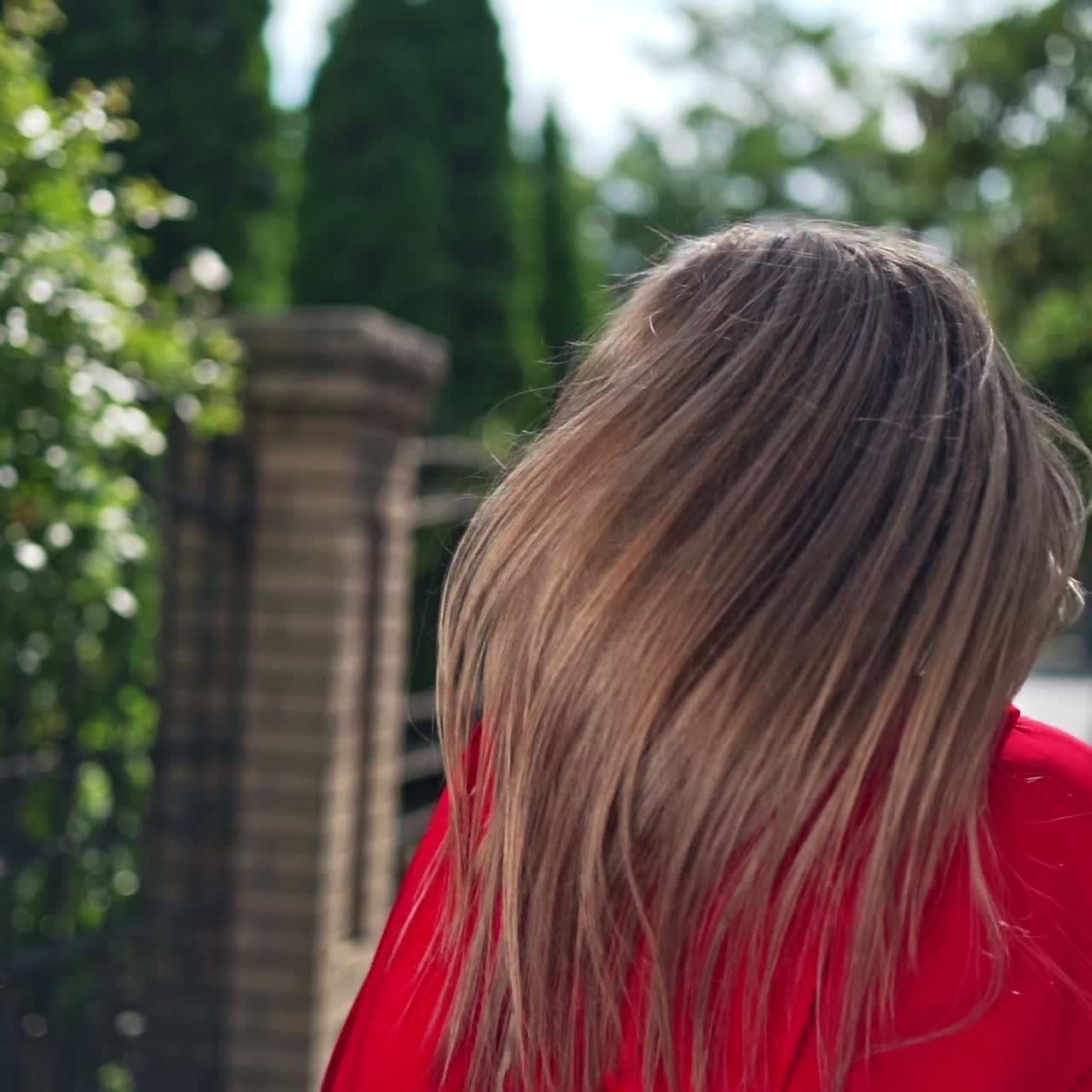 Passionate young girl posing to camera. Beautiful young model in red dress with long hair on the green trees background. Sexy woman turning around herself joyfully.