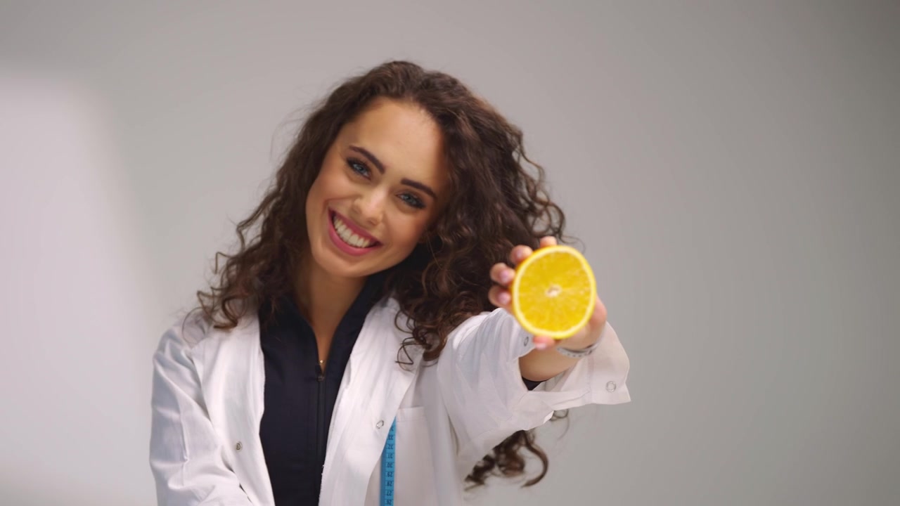 Young female dietitian holding sliced orange toward camera and smiling in white studio. Healthy eating, fresh fruit, wellness lifestyle and nutrition guidance concept.