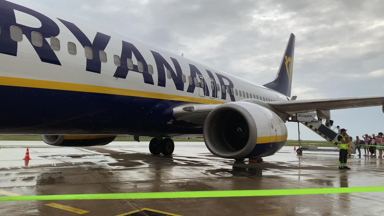 Passengers board an aircraft under grey, rainy skies, stepping carefully as raindrops fall, reflecting on the wet tarmac, adding a calm yet rushed atmosphere.