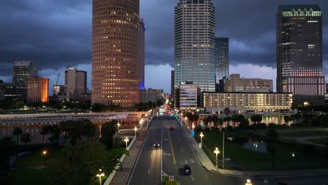 tampa’s city skyline rises over a quiet street at dusk, featuring rivergate tower and high-rise buildings. cars move below illuminated streetlights on a major road. aerial view.