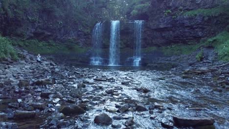 Drone-Volando-Más-Allá-De-Una-Chica-Sentada-En-Las-Rocas-Al-Lado-De-Una-Cascada