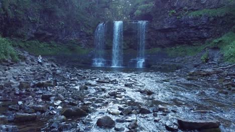 drone flying past a girl sitting on the rocks next to a waterfall