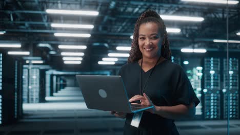 retrato de una exitosa mujer afroamericana especialista en ti que usa una computadora portátil, de pie en un centro de datos. el administrador de sistemas trabaja en servicios web. computación en la nube, análisis de servidores, seguridad cibernética