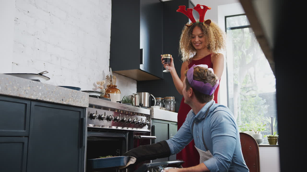 Coppia con corna da renna e cappello di carta a casa, che cucina una cena vegetariana il giorno di Natale.