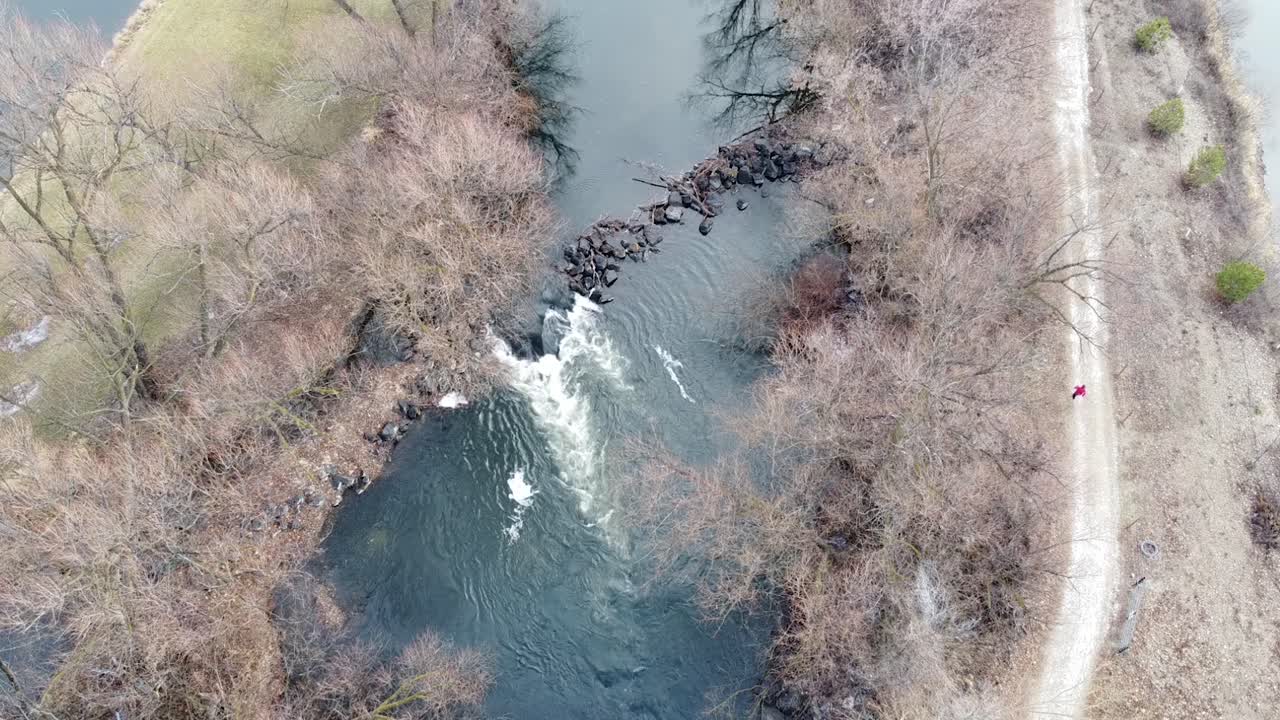 Small river dam and rapids on scenic, idyllic, willow tree lined river in winter with a person wearing red running on the track in rural countryside of Boise, Idaho, USA
