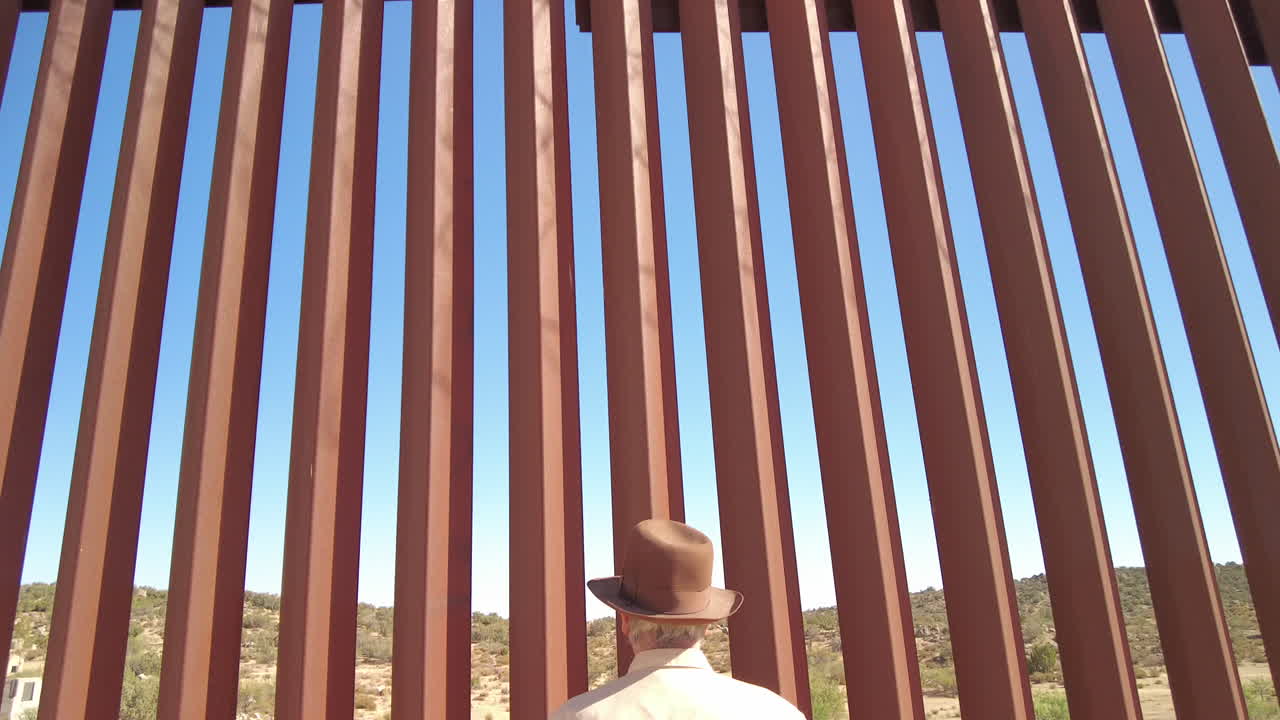 Mature caucasian American male with hat looks through gaps of US Mexico border fence in Jacumba, tilt down shot