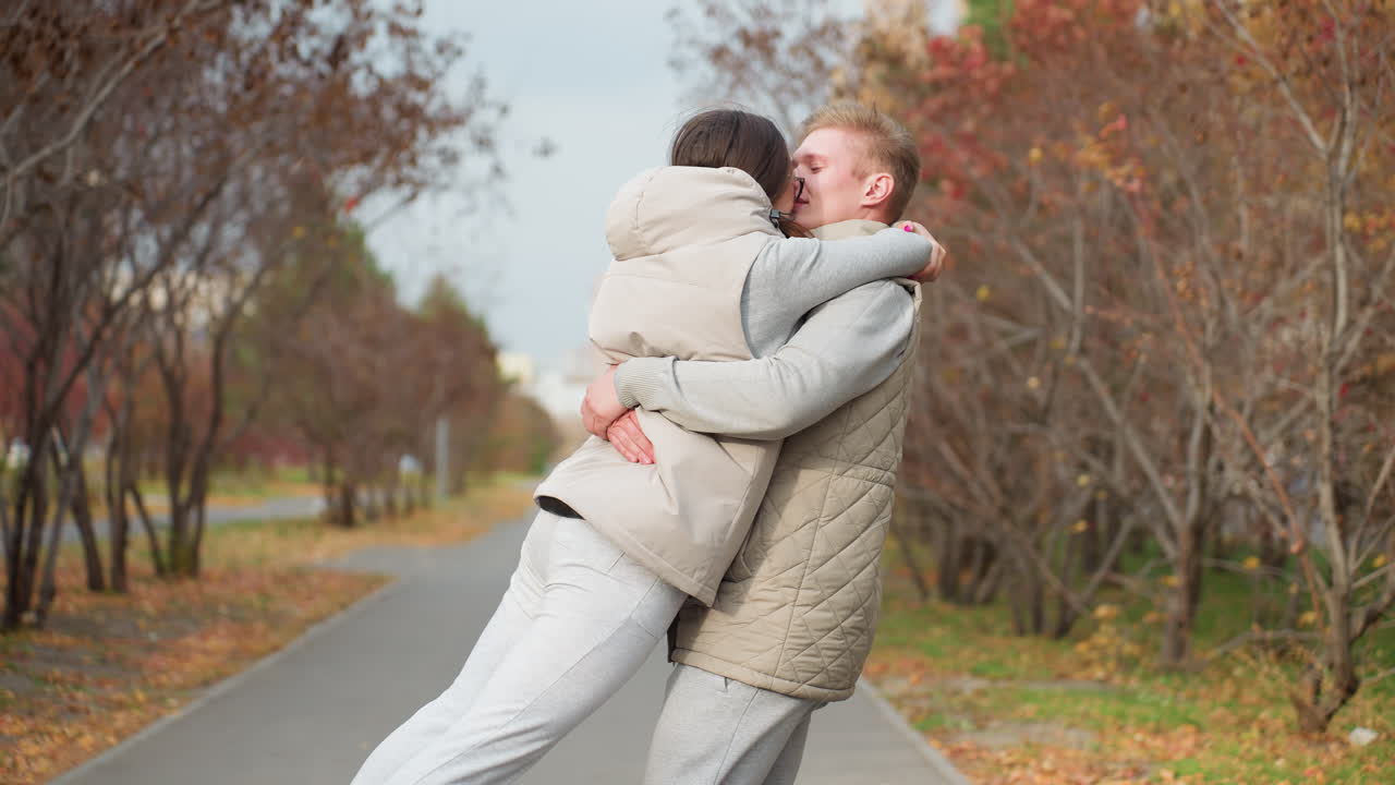 Newly married adult couple hug affectionately as man lifts wife up and spins her around joyfully, sharing tender kiss outdoors on peaceful park pathway with trees and autumn leaves all around them