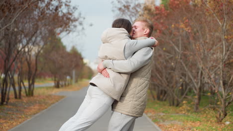 newly married adult couple hug affectionately as man lifts wife up and spins her around joyfully, sharing tender kiss outdoors on peaceful park pathway with trees and autumn leaves all around them