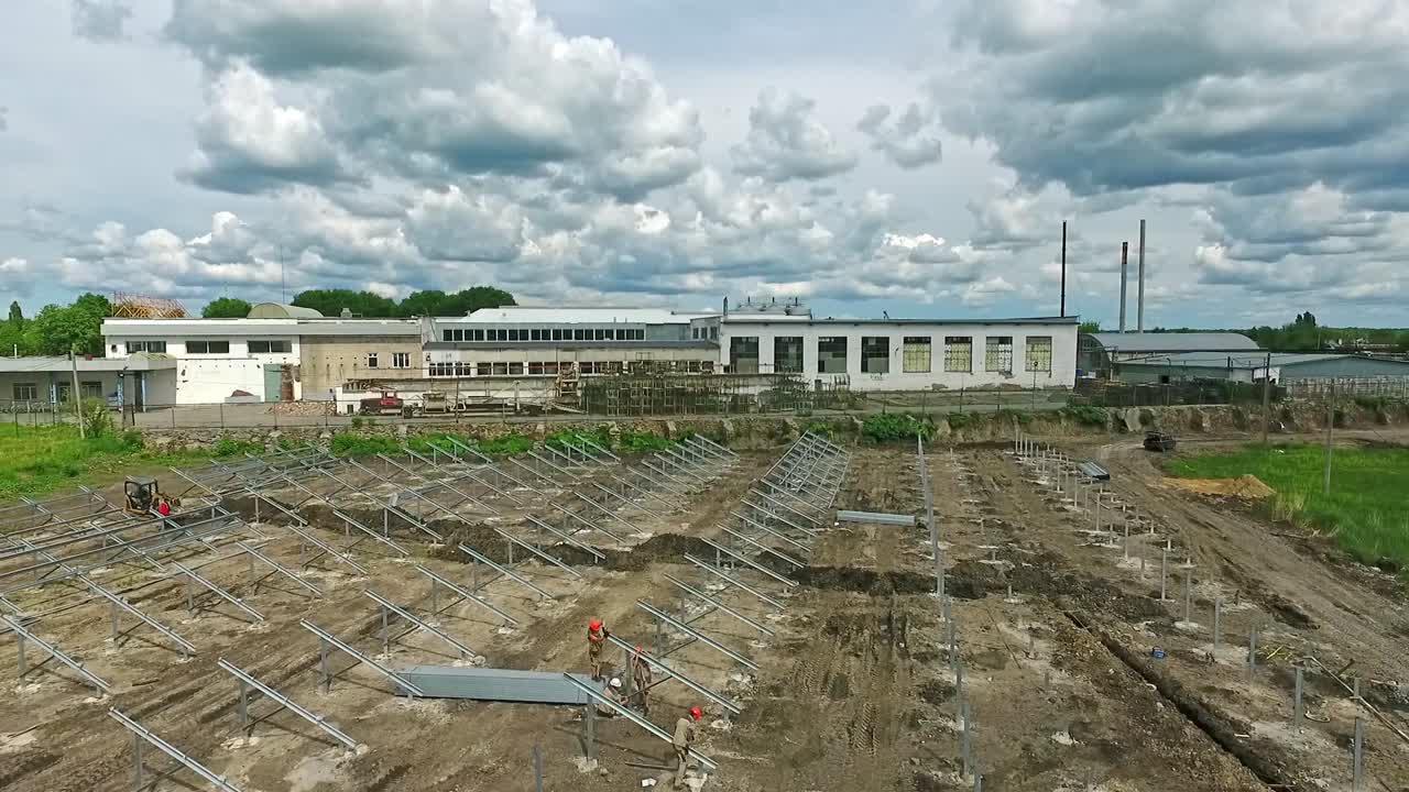 Construction site with workers working on. Metal supports prepared on the ground for installing solar panels. Industrial warehouses at backdrop.