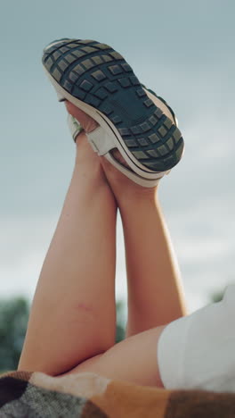 vista lateral de una mujer con pantalones cortos acostada en una manta con las piernas levantadas y cruzadas en sandalias, el cielo nublado y el fondo borroso crean una atmósfera al aire libre pacífica