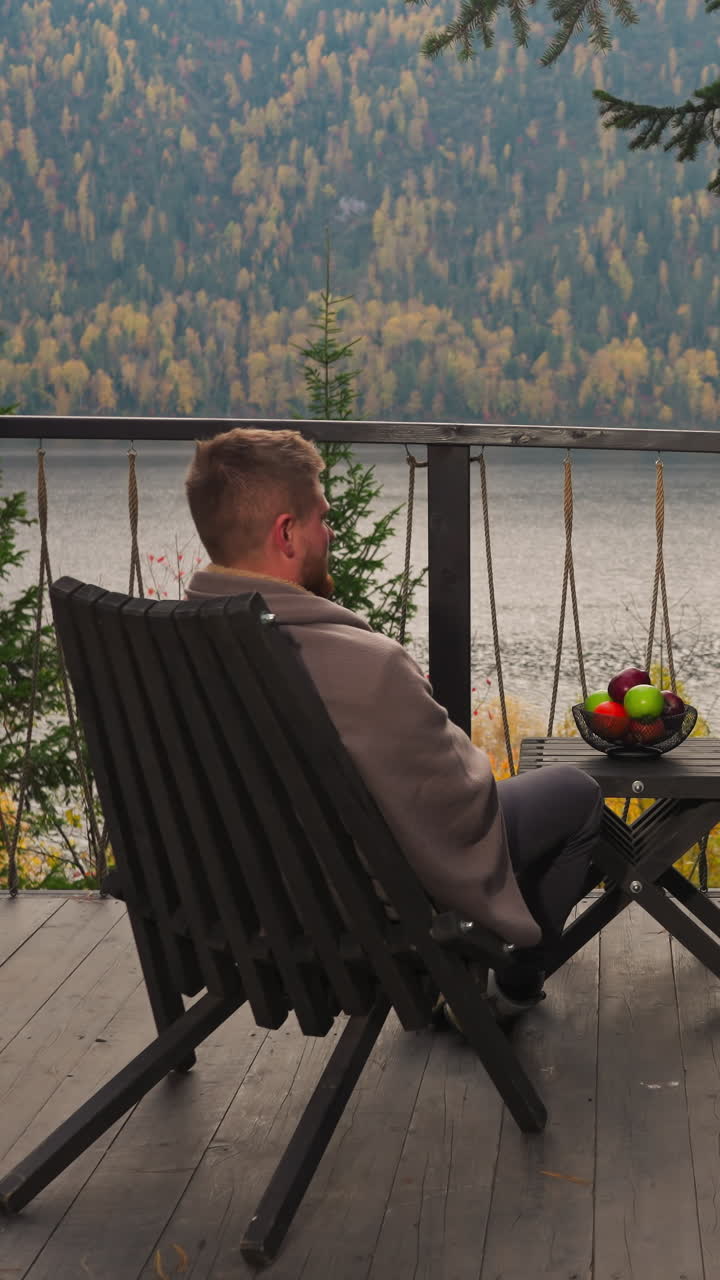 Woman and man on terrace sit at table with fruits. Friends enjoy time together on balcony against river and mountains. Ideal autumn time-spending