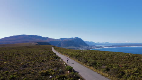 Cyclist-on-scenic-mountain-road-amongst-fynbos-with-epic-ocean-views,-Hermanus