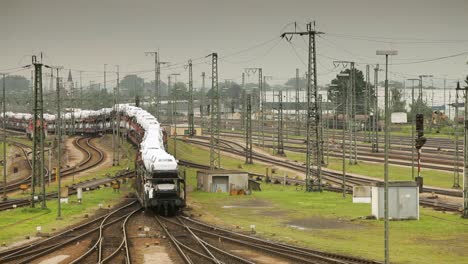 cámara de mano captura un tren de carga blanco con una franja azul curva alrededor de una curva en medio de un patio de trenes ocupado, múltiples vías y trenes, postes eléctricos bajo un cielo nublado