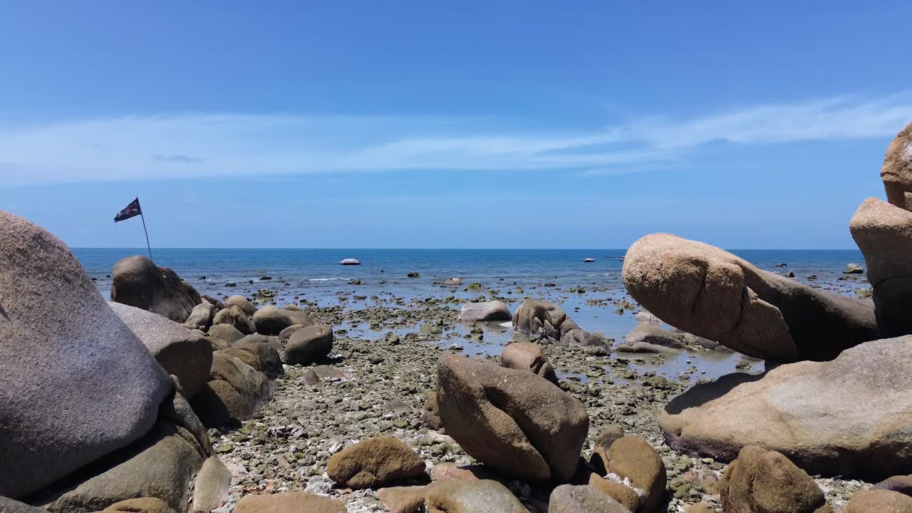 A pirate flag waves on a rocky shoreline at Lamai Beach, Koh Samui, Thailand. Turquoise water, clear blue sky, and scenic coastal landscape