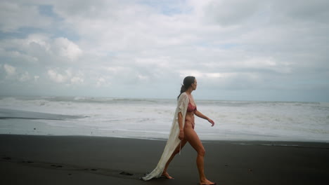 attractive, young, independent woman in bikini walks on black sand beach alone in bali, indonesia