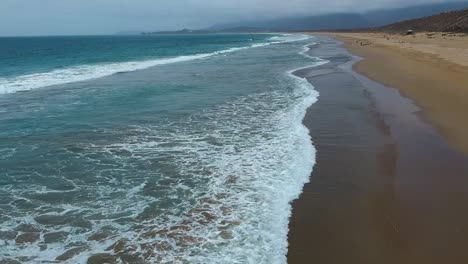 De-Un-Lado-El-Océano-Del-Otro-La-Arena-Toma-Aérea-Moviéndose-Hacia-Adelante-A-Lo-Largo-De-Las-Olas