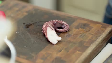 Close-up-of-male-hands-cutting-the-initial-part-of-a-fresh-purple-octopus-tentacle,-with-a-kitchen-knife,-on-a-wooden-cutting-board,-while-someone-is-explaining-what-to-do,-in-a-professional-kitchen