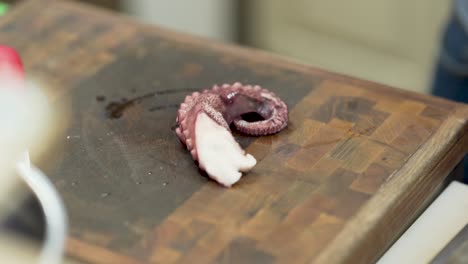 cierre de manos masculinas cortando la parte inicial de un tentáculo de pulpo morado fresco, con un cuchillo de cocina, en una tabla de corte de madera, mientras alguien explica qué hacer, en una cocina profesional