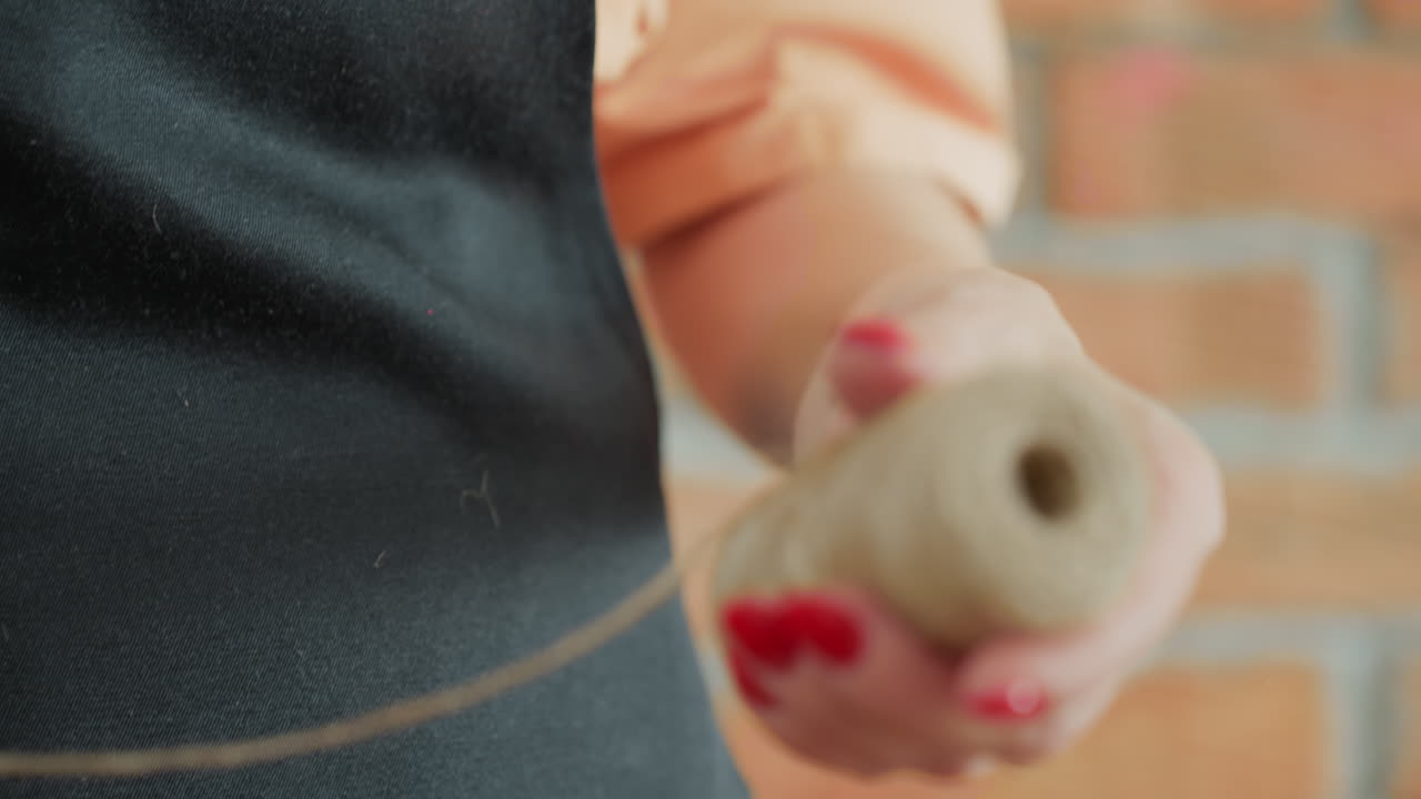 Decorator in apron holding spool of natural twine, pulling rustic string with careful hands, preparing material for creative handmade decoration project in cozy workshop with artisan atmosphere