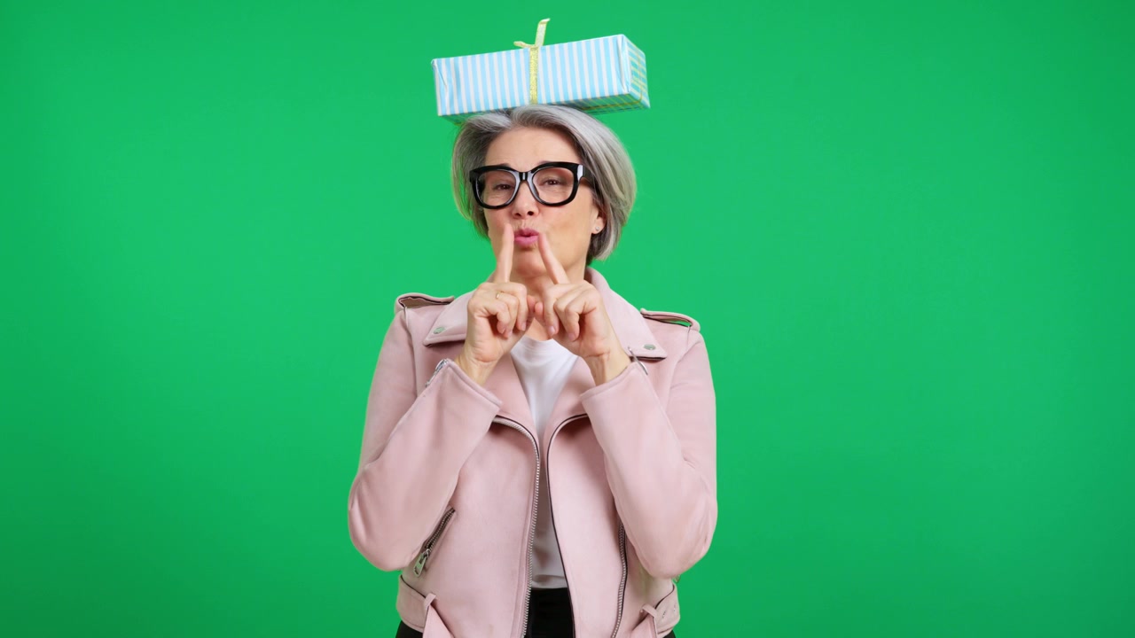Elegante donna matura che bilancia una scatola regalo sulla testa davanti a uno schermo verde