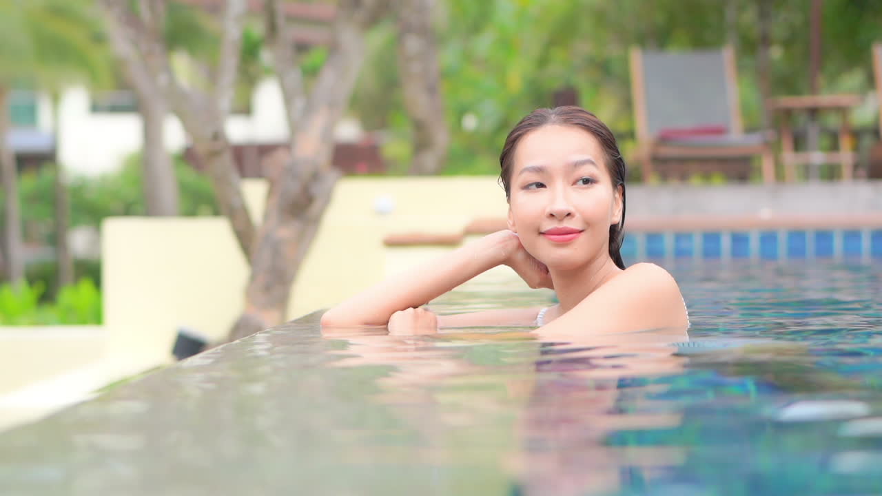 열대 휴가 동안 인피니티 풀 가장자리에서 팔꿈치에 기대어 옆으로 카메라를 바라보며 쉬고 있는 여성