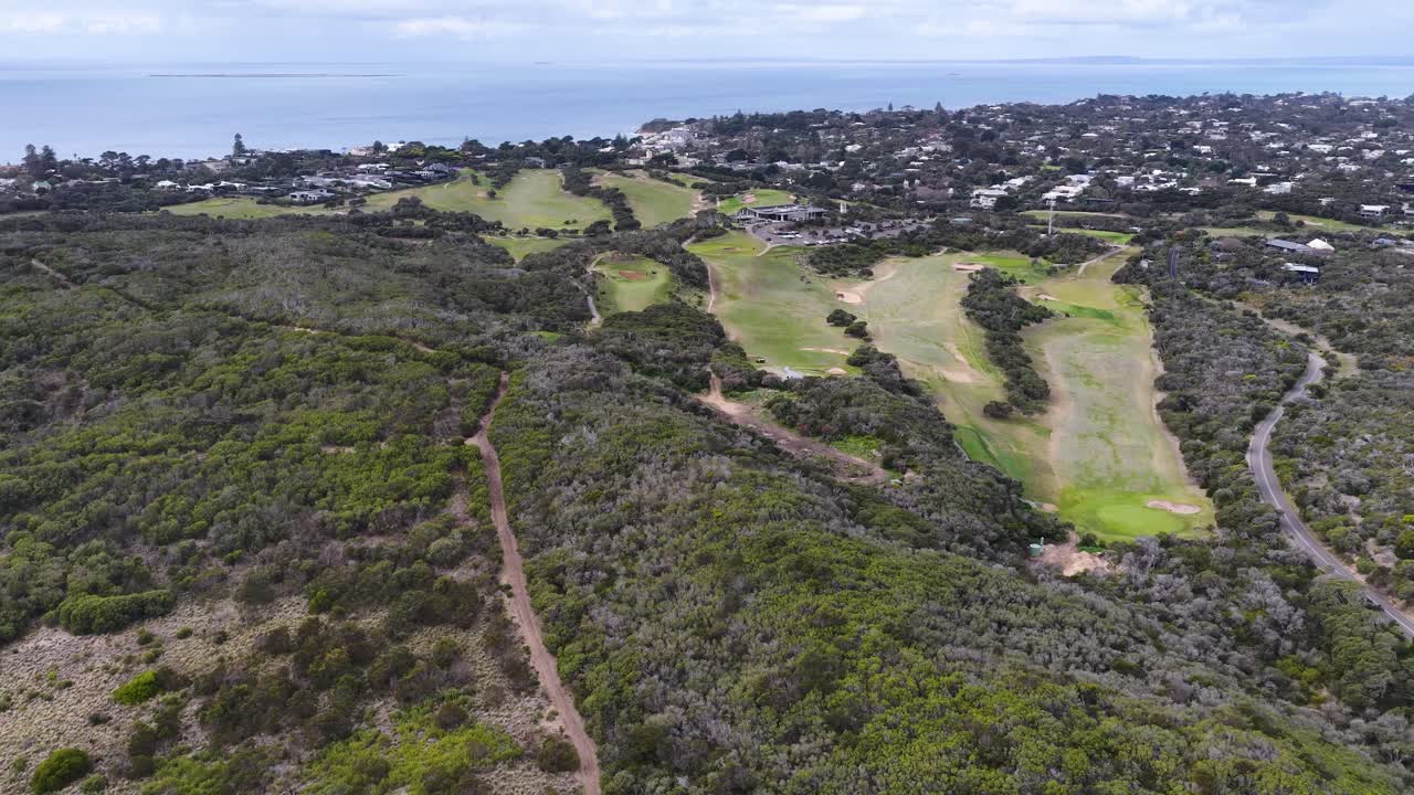 Drone glides above lush golf course, coastal vegetation, and residential area under soft daylight