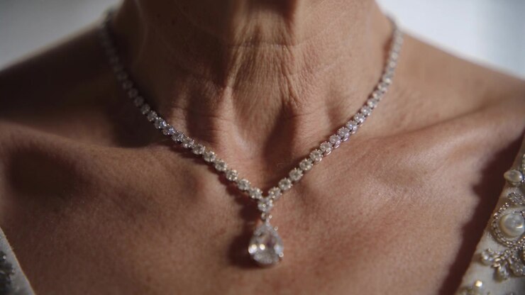 Mostrando una donna anziana che respira, il collo che si muove in una luce soffusa, un abito con perline che incornicia un ciondolo di diamanti.