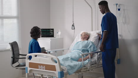 a team of african ethnic group doctors a man and a woman talk to an elderly male patient. a doctor and a nurse talk to a bedridden patient in a hospital ward