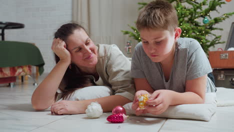 boy in casual wear lying on marble floor holding colorful christmas ornament focused while mother lying beside him smiling warmly decorated christmas tree background cozy indoor