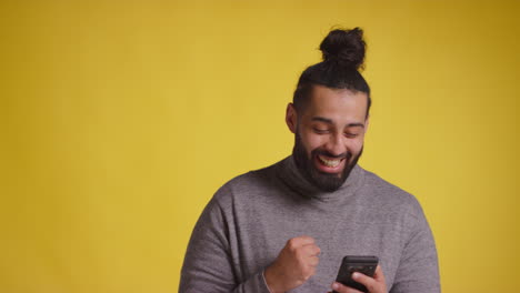 Fotografía-De-Estudio-De-Un-Joven-Sonriente-Con-Un-Teléfono-Móvil-Recibiendo-Buenas-Noticias-Contra-Un-Fondo-Amarillo-1