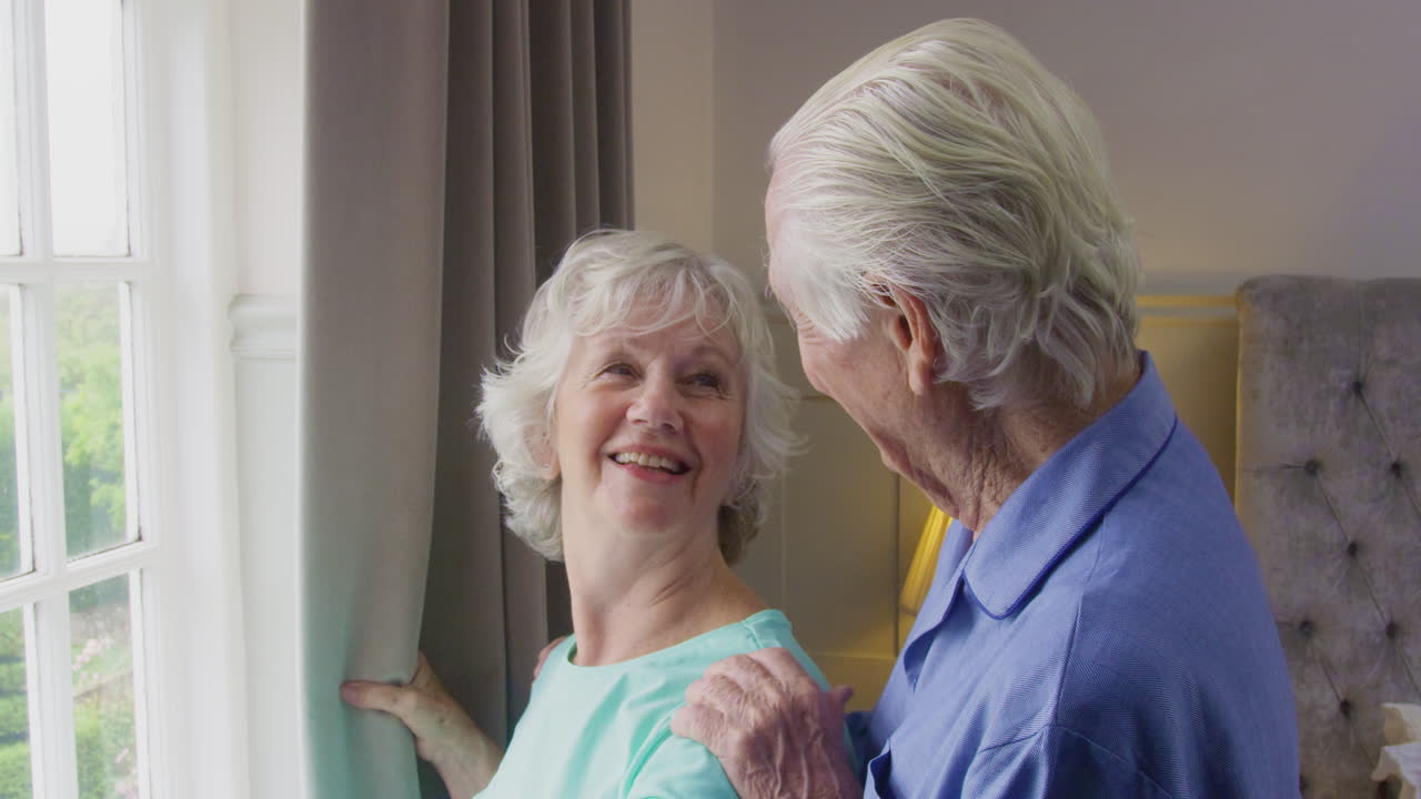 잠옷을 입고 침실 커튼을 열고 창밖을 바라보는 미소 짓는 고령 부부