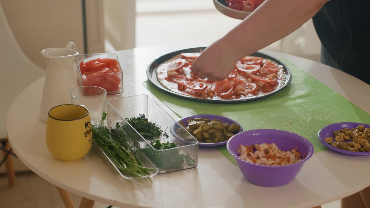 Mani che posizionano pomodori a fette su impasto per pizza coperto di salsa su un vassoio metallico rotondo, mentre ciotole di formaggio grattugiato, olive, erbe tritate, cipolle e utensili da cucina riposano su un tavolo bianco accanto a una finestra con