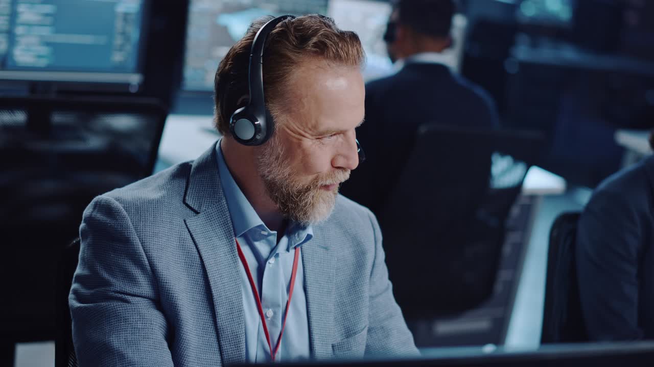Joyful Bearded Senior Technical Support Specialist is Talking on a Headset while Working on a Computer in a Dark Monitoring and Control Room Filled with Computer Display Screens and Data Servers.