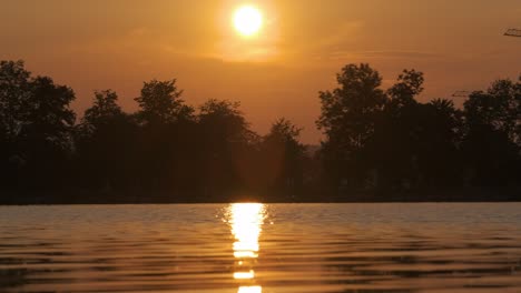 paisaje a orillas del lago con silueta oscura de árboles del parque reflejados en el agua del lago y gente peatonal caminando lejos en el terraplén al atardecer brillante