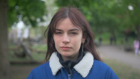 Brünette-Frau-In-Jeansjacke-Blickt-In-Zeitlupe-In-Einem-Park-Ernsthaft-In-Die-Kamera