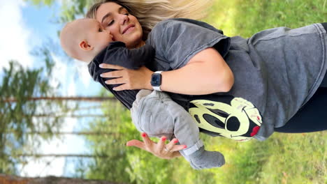 blonde long-haired woman wearing sportive clothes holding her baby. mother embracing her little son in nature. vertical screen.