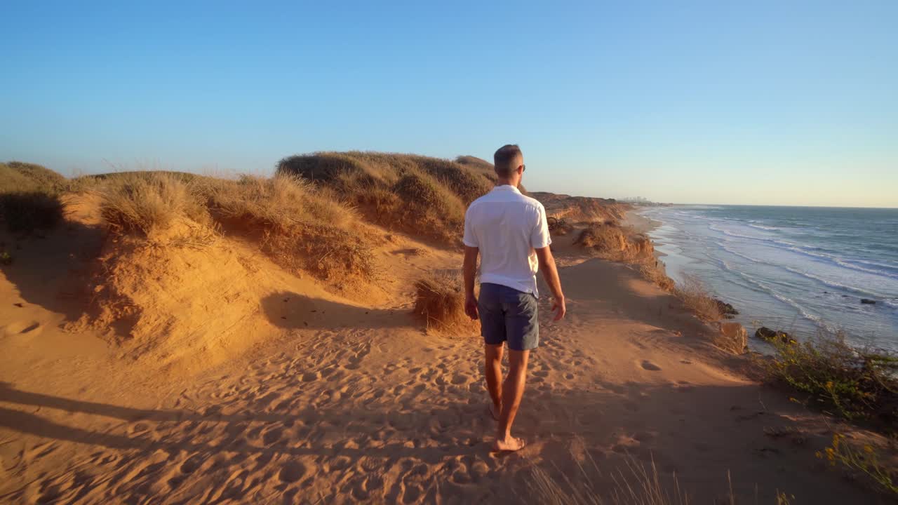 un hombre solitario con camiseta blanca y medio pantalón caminando en una playa dorada al atardecer en haderah, israel