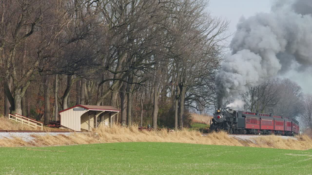 A classic steam train chugs along the tracks surrounded by lush trees and fields, evoking nostalgia in a peaceful rural setting during autumn.