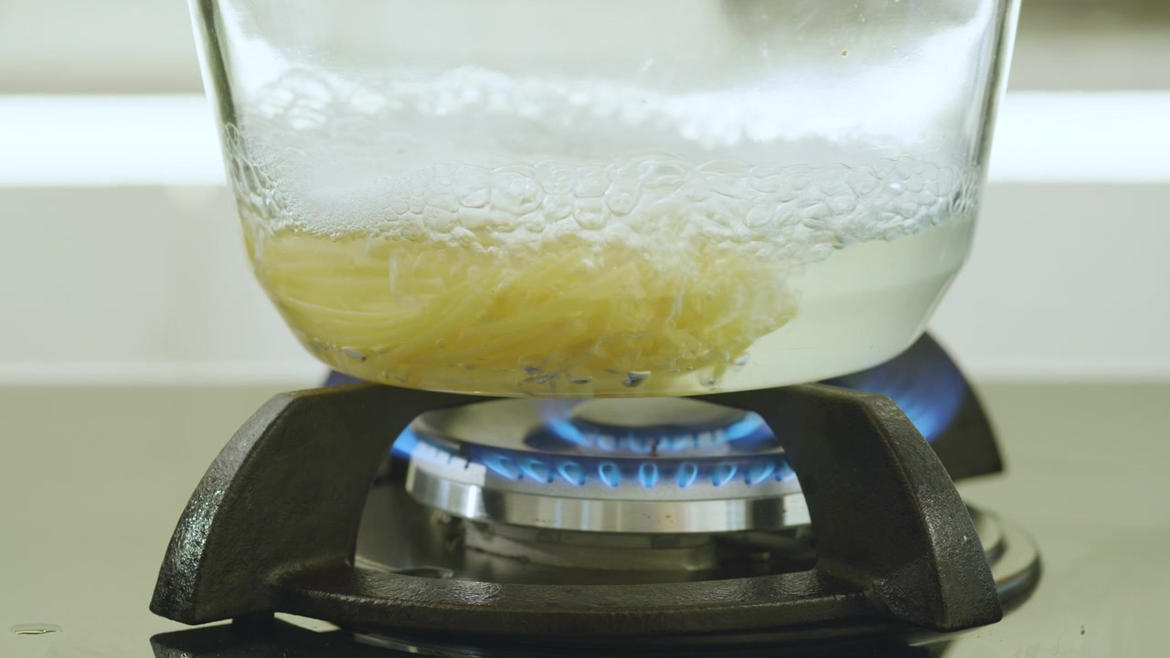 Primo piano che mostra un contenitore di vetro trasparente con liquido bollente e noodles, poggiato su una griglia del fornello con una fiamma blu visibile.