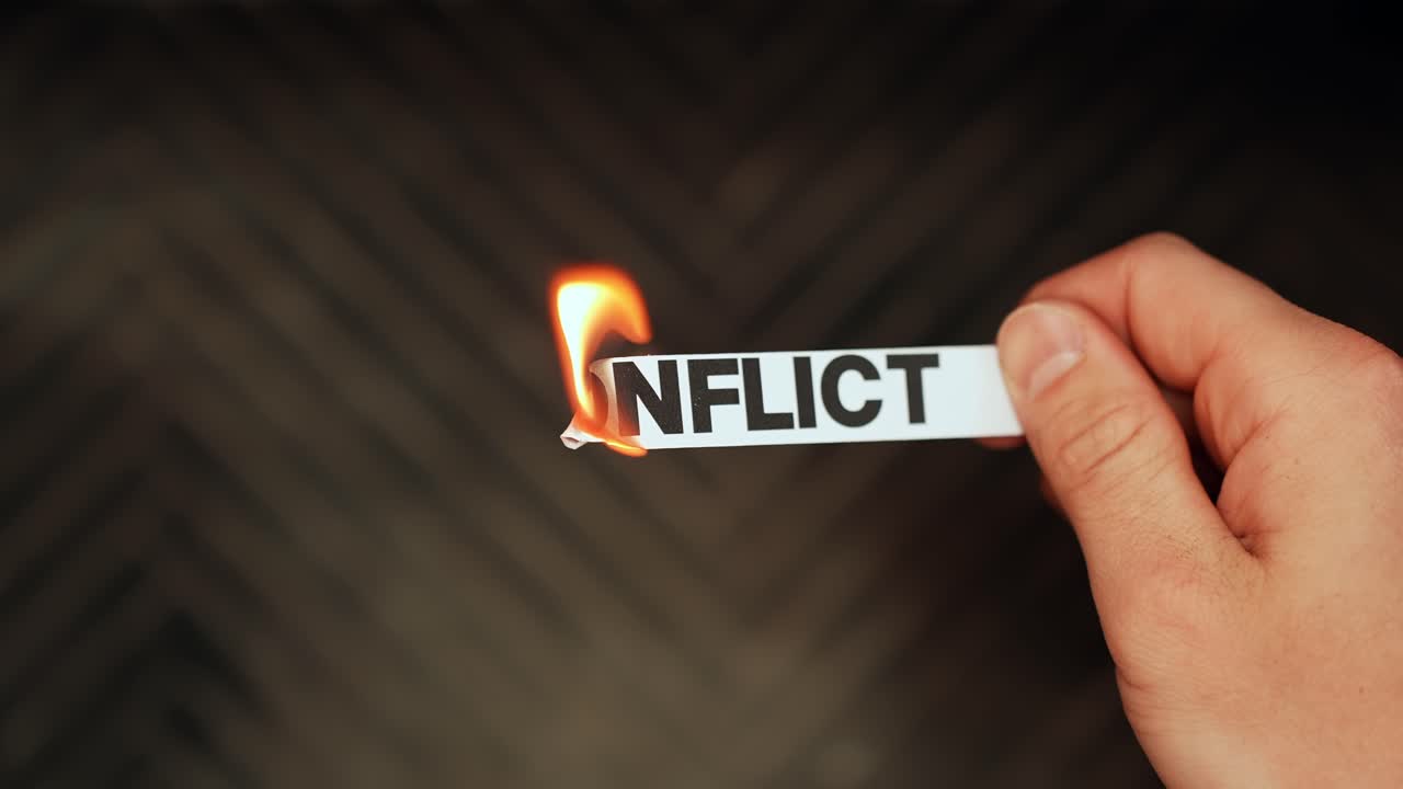Close-up of a “CONFLICT” paper strip being held while the edge ignites, symbolizing dispute, tension, and crisis on a dark background with copy space