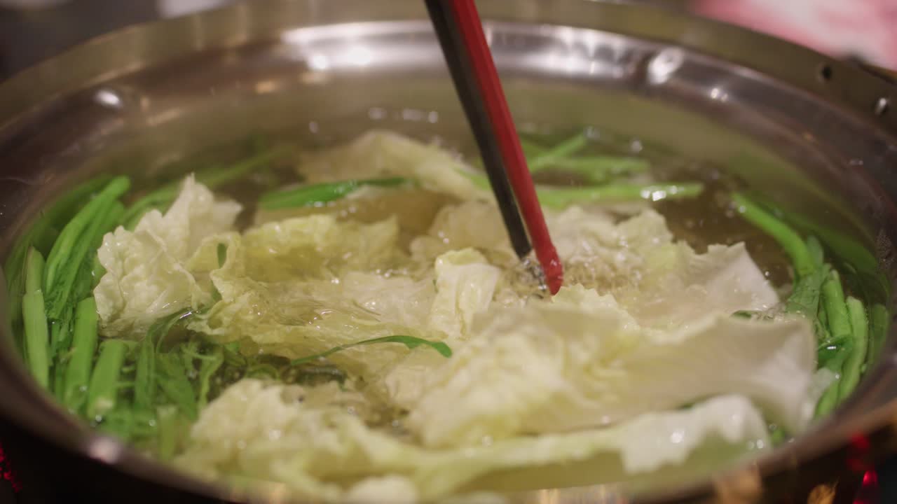 Fresh napa cabbage placed in simmering hotpot broth with greens, stirred gently using chopsticks