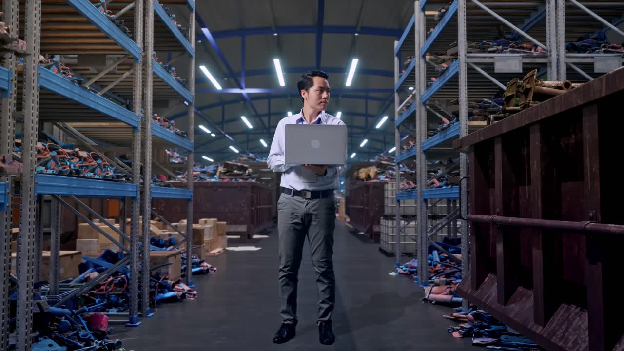 Figura intera di un professionista uomo asiatico con il suo laptop, in un magazzino industriale di rottami metallici e riciclaggio. Osserva l'ambiente circostante prima di concentrarsi sul suo laptop.