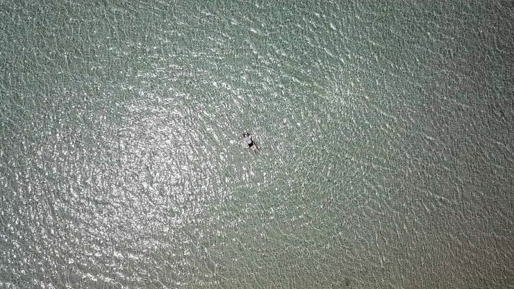 AERIAL RISING shot from a teenage girl lying in shallow water on a beach