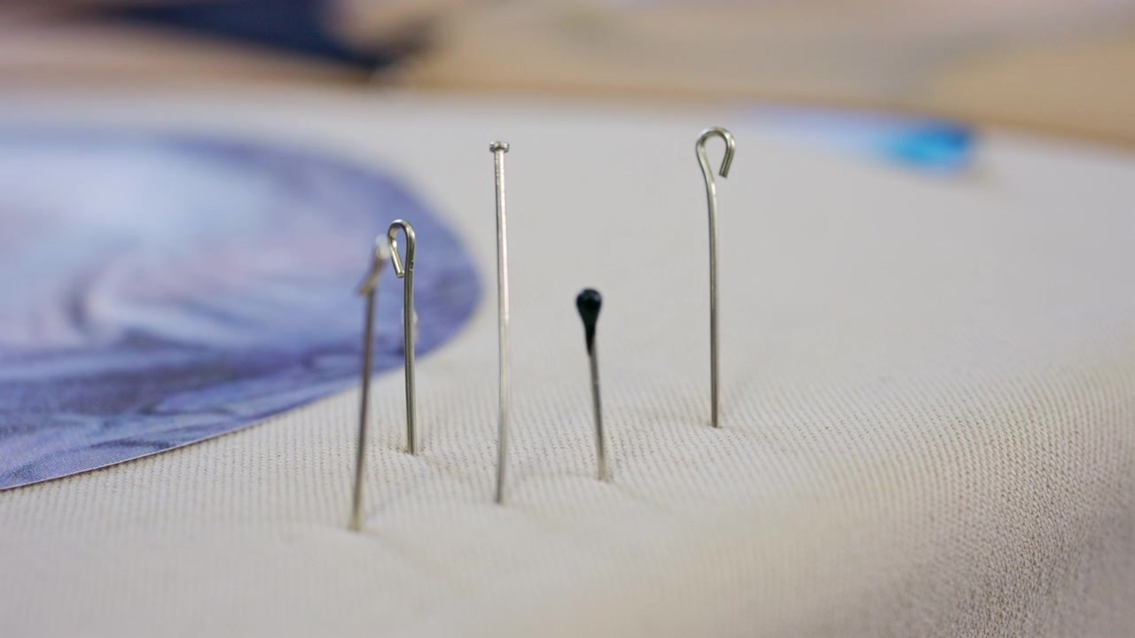 Macro shot of two thin metal sewing pins standing upright in beige woven fabric, with a soft blue paper or fabric pattern blurred in the background