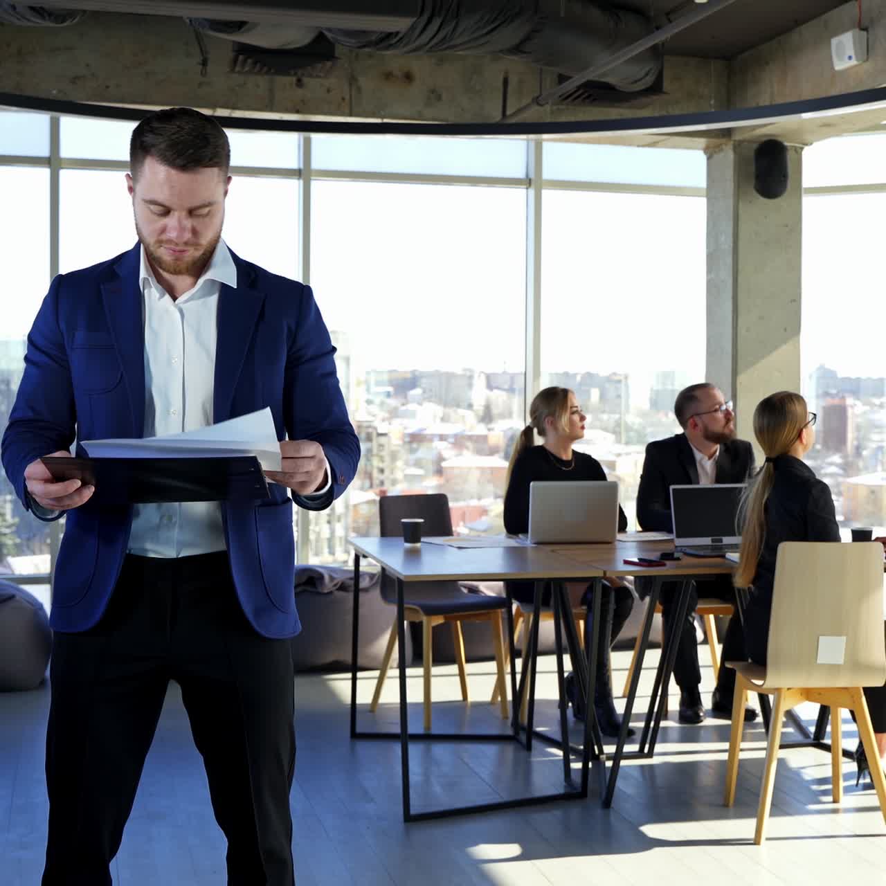 Serious entrepreneur in suit in office room. Business people planning new blueprint together. Businessman stands alone holding folder with papers